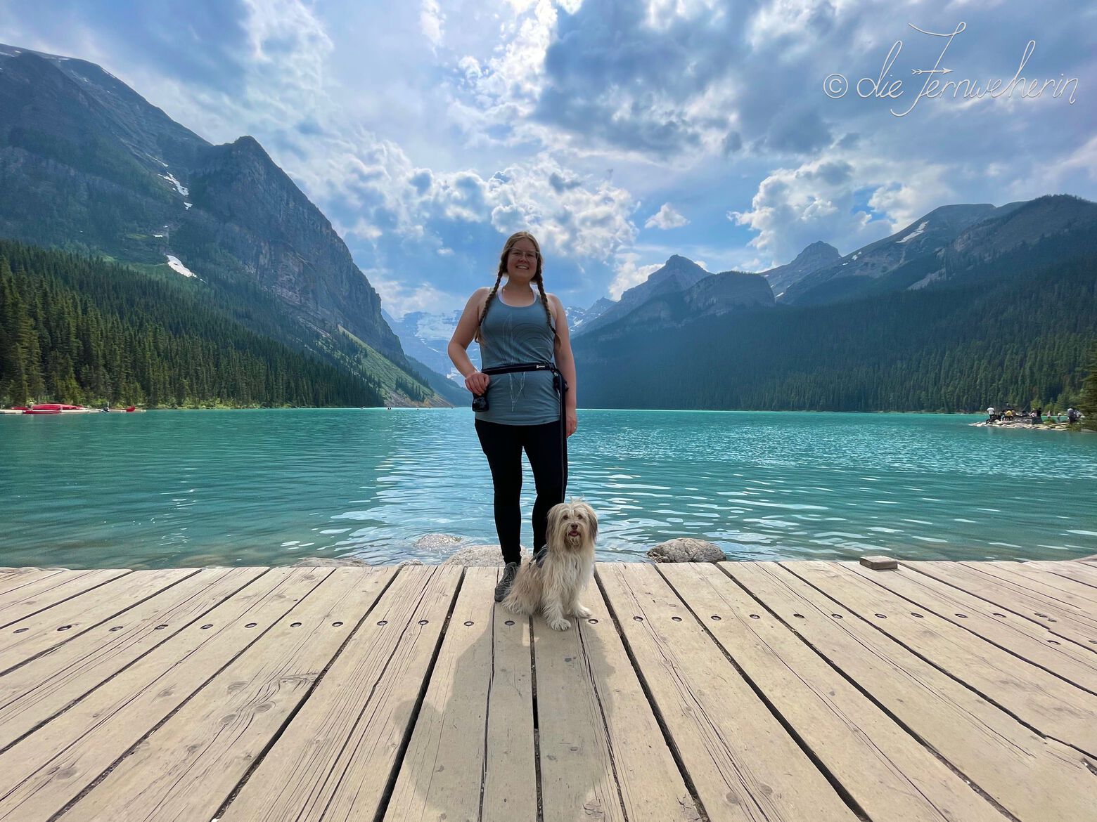 A woman and a dog pose on the boardwalk in front of beautiful Lake Louise in Banff National Park.
