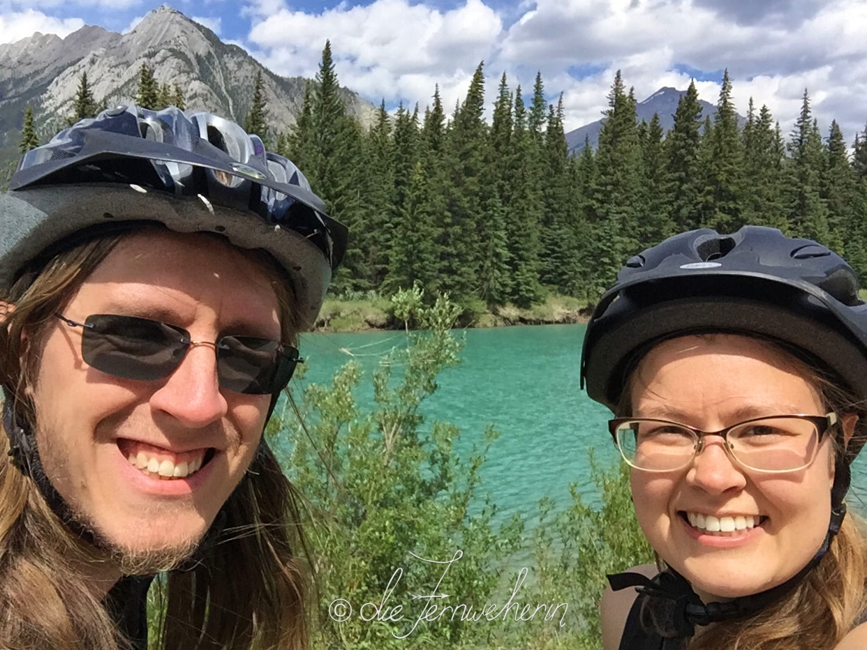 Two cyclists wearing helmets pose in front of Bow River on the Sundance Trail in the town of Banff.
